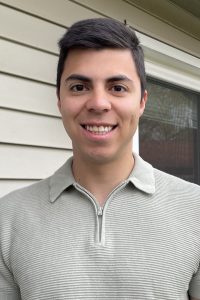 An image of Alejandro Casco, a man with dark hair and dark eyes, smiling in front of a house wearing a tan 3/4 zip.